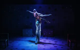 A male dancer lifts a female dancer on a dark stage.