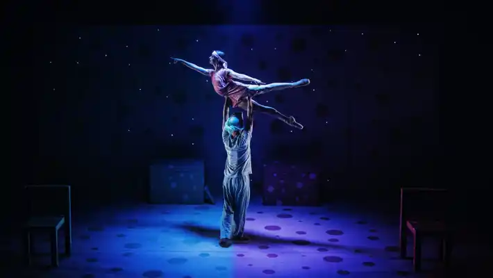 A male dancer lifts a female dancer on a dark stage.