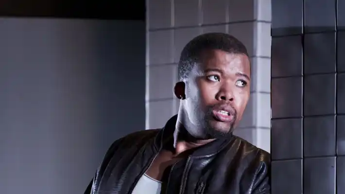 A short-haired man in a leather jacket leans against a tiled wall.