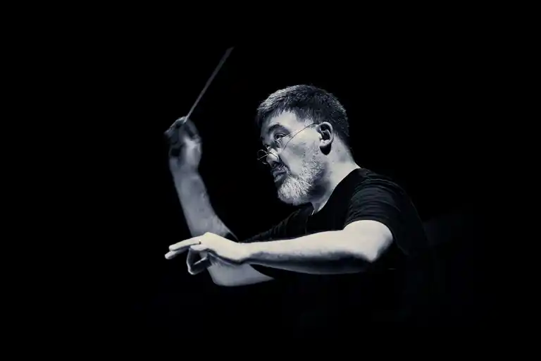 A man in a black short-sleeved shirt with glasses and a conductor's baton in his hand against a dark background.