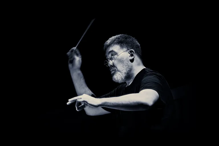 A man in a black short-sleeved shirt with glasses and a conductor's baton in his hand against a dark background.