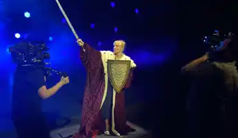 On a stage with blue lights, a woman stands with a shield and sword, two men with cameras are filming.