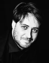 A close-up of a man with a beard against a dark background.