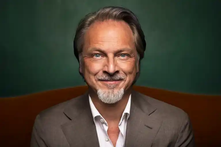A smiling person in a grey suit and white shirt in front of a green wall.