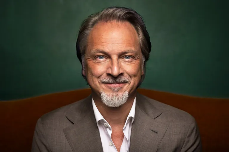 A smiling person in a grey suit and white shirt in front of a green wall.