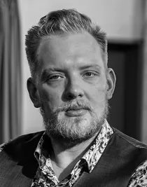 Portrait image of a blond man with a beard and mustache, he is wearing a vest and a floral shirt.