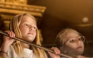 Two people with blurred faces stand behind a railing indoors, surrounded by warm tones.