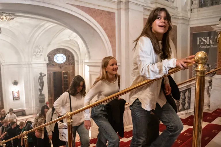 People walk on a staircase in an elegant building with arches and chandeliers.