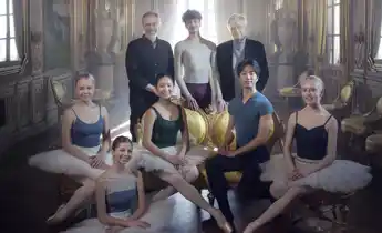Six young dancers flanked by two older gentlemen in a light-filled room.