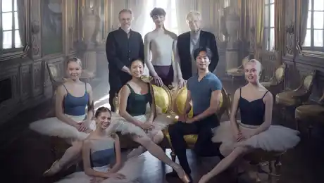 Six young dancers flanked by two older gentlemen in a light-filled room.