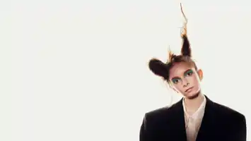 En ung kvinna, iförd mörk kavaj och med håret uppsatt i två koner, huvudet lätt lutat åt sidan, mot en vit bakgrund.