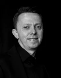 A close-up of a smiling man, dressed in dark clothes, against a dark background.