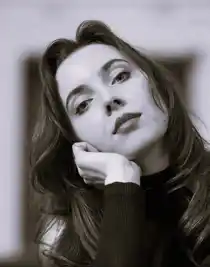 A close-up of a woman with long dark hair, she tilts her head to the left and rests it in her hand.