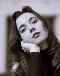 A close-up of a woman with long dark hair, she tilts her head to the left and rests it in her hand.