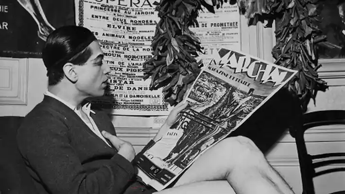 A barefoot man sits in profile and reads a newspaper.