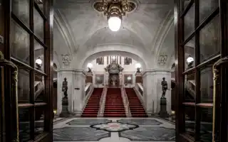 View of a large entrance hall, a staircase with red carpet leads up to a door.
