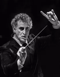 A man in a suit raising both hands, in one hand a conductor's baton.