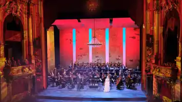 A beautifully lit stage, surrounded by gold decor, with an orchestra and singers in the foreground.