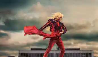 A figure in a red robe stands in front of a dramatic sky, with a red cloth dancing in the wind and the silhouette of a house in the background.