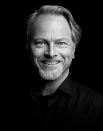 A close-up of a smiling man with a beard in a dark shirt.