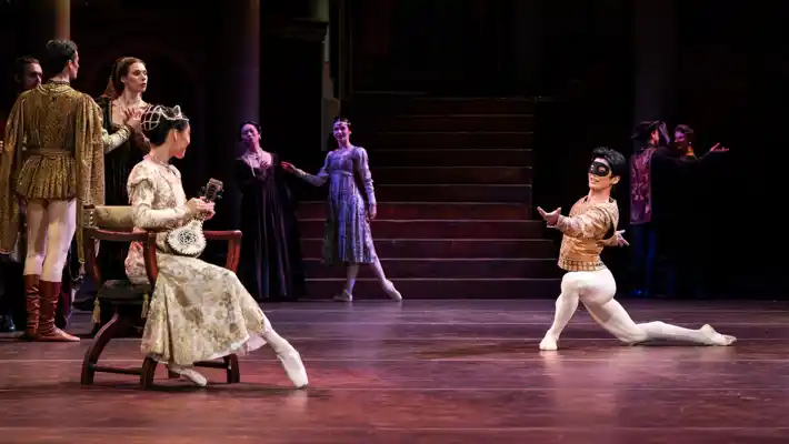 Several dancers on a stage, a man kneels in front of a woman sitting in a chair.