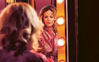 A woman sits with her back to us, she looks into a mirror surrounded by lights.