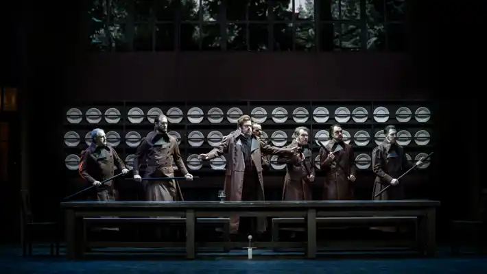 Several people are standing at a large table, behind them a wall adorned with plates on racks.