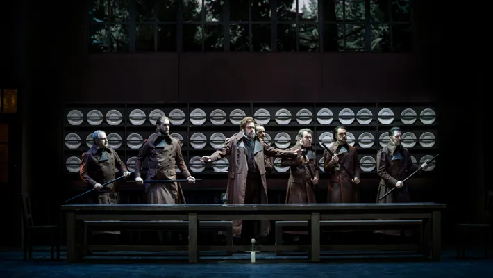 Several people are standing at a large table, behind them a wall adorned with plates on racks.
