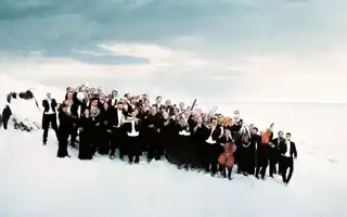 A group of people in black suits, some with instruments, stand in a winter landscape.