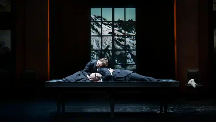 A woman and a man are half-reclining on a large table, in the background a window overlooking an Alpine landscape.