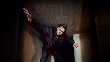 A person in dark clothes strikes a dramatic pose in a narrow concrete corridor.