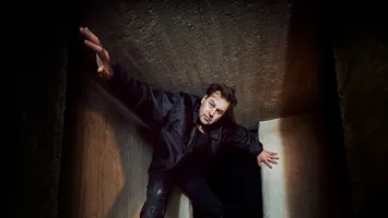 A person in dark clothes strikes a dramatic pose in a narrow concrete corridor.