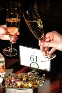 Två personer skålar med champagne i glas, på bordet står olika snacks.