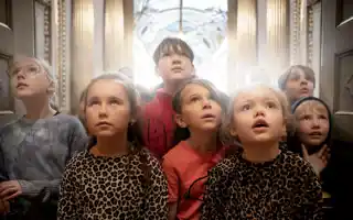 Group of children in different clothes standing together, indoors with gold columns in the background.