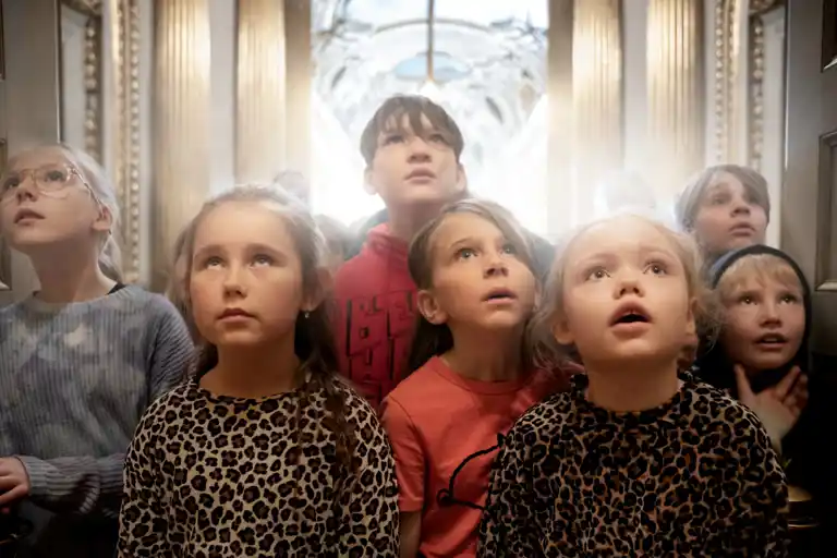 Group of children in different clothes standing together, indoors with gold columns in the background.