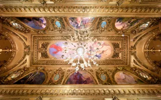 View of a richly decorated ceiling with gold and paintings, in the center a large chandelier.