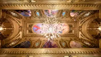 View of a richly decorated ceiling with gold and paintings, in the center a large chandelier.