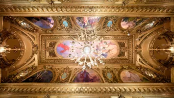 View of a richly decorated ceiling with gold and paintings, in the center a large chandelier.