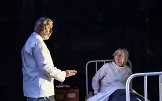 A woman is sitting in a hospital bed, a man in what looks like hospital clothes is talking to her.