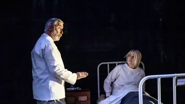 A woman is sitting in a hospital bed, a man in what looks like hospital clothes is talking to her.