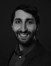 Close-up of a smiling man with dark hair, beard and mustache; he is wearing a dark shirt and jacket.