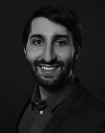Close-up of a smiling man with dark hair, beard and mustache; he is wearing a dark shirt and jacket.