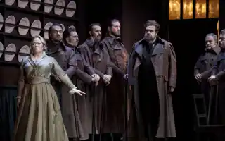A group of people stand in a kitchen-like environment, plates lined up on the wall behind them.