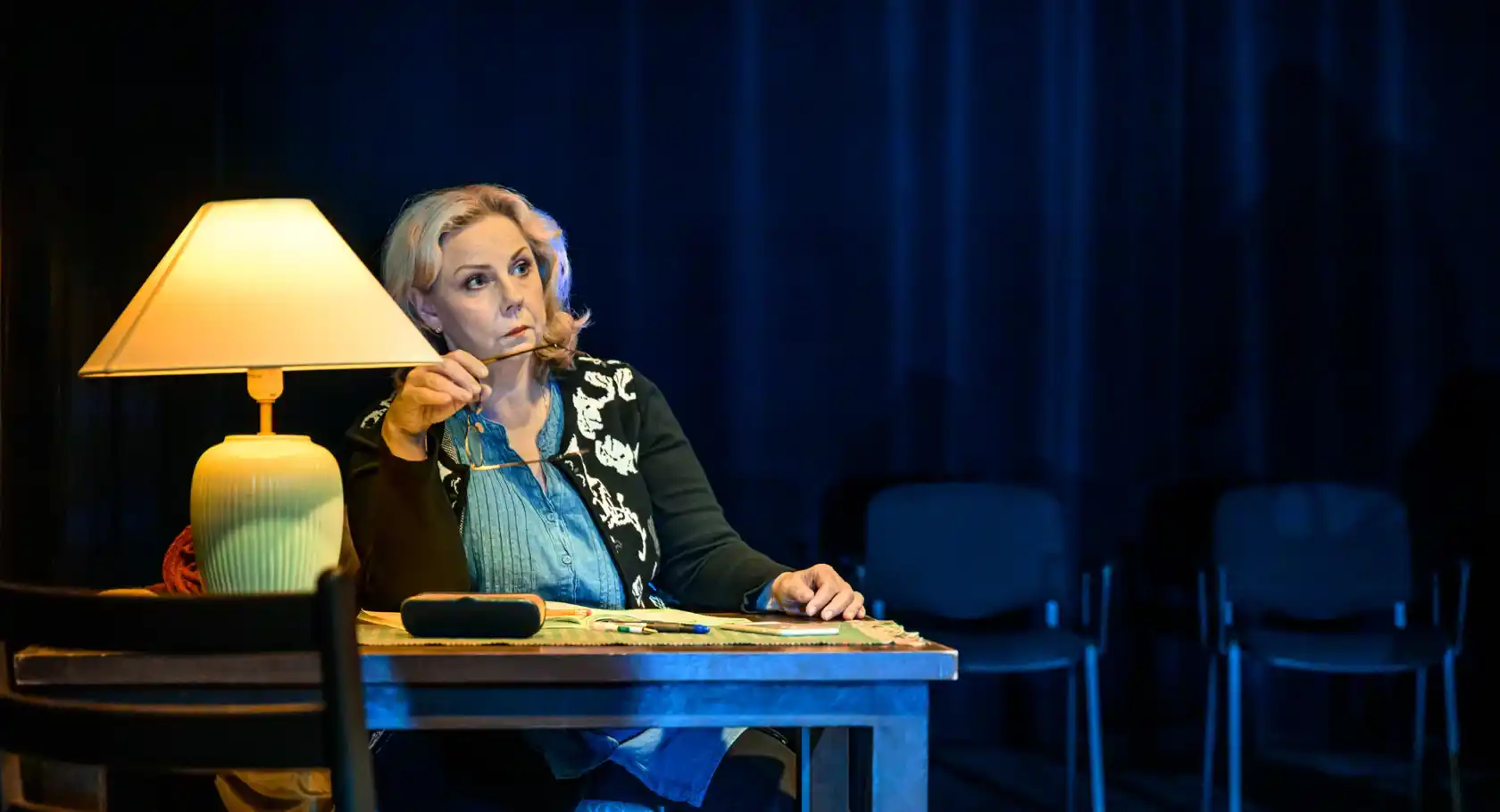 A woman, holding a pair of glasses, sits at a desk, on the table stands a lamp.