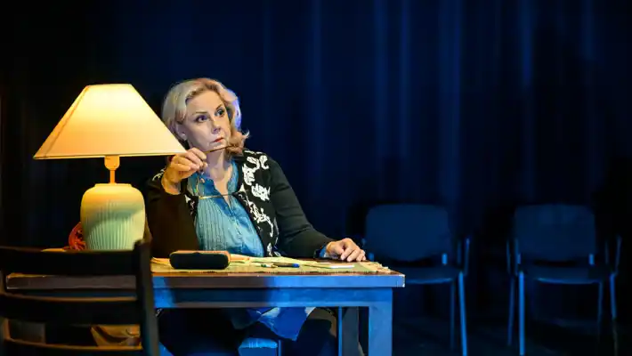 A woman, holding a pair of glasses, sits at a desk, on the table stands a lamp.