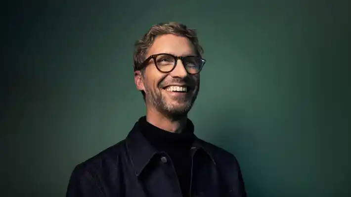 En skrattande man i glasögon framför en mörkt grön bakgrund.