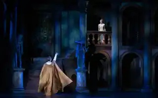 A man, with an arm stretched out, is standing below a balcony, on the balcony stands a young woman.