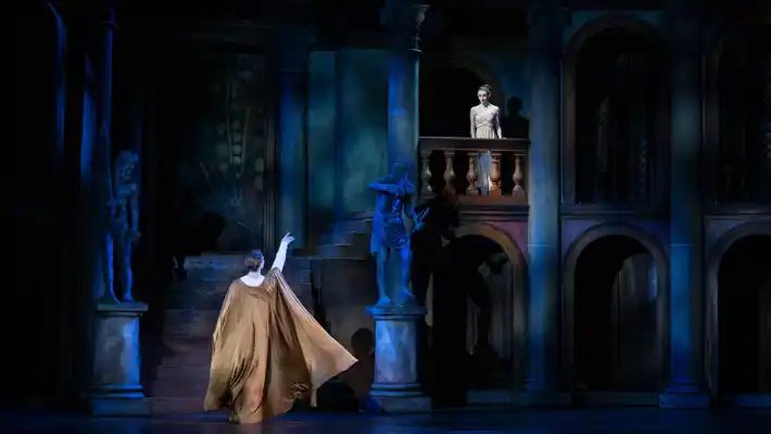 A man, with an arm stretched out, is standing below a balcony, on the balcony stands a young woman.